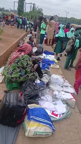 The video shows an outdoor market scene. In the foreground, numerous items are displayed on the ground, including clothing, shoes, and bags. Several people are present, some appearing to be vendors and others potential customers. In the background, there are more people, motorcycles, and what appears to be a street or road. The overall impression is a busy, informal marketplace.