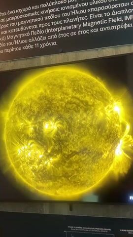 A close-up, medium-angle shot shows a highly detailed, bright yellow image of the sun, with numerous flares and prominences visible. The image is displayed on a screen or as a print. Above the sun image, text in what appears to be Greek is visible on a dark background. The lower part of the image shows a dark surface, possibly a display stand or railing, with more text below that, also appearing to be in Greek. The overall scene is dimly lit except for the bright sun image.