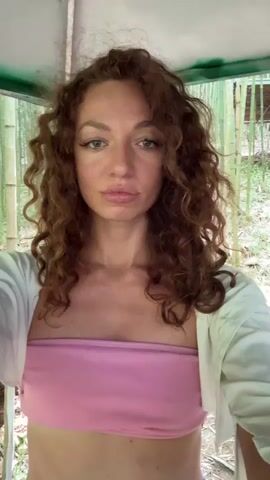 The video shows a woman in a pink tube top and white shirt standing under a covered structure. Behind her is a man in a green t-shirt and baseball cap looking at his phone. Outside the structure, there's a dense growth of bamboo. The woman appears to be recording the video. She looks at the camera, then gestures slightly to the man, then looks back at the camera.