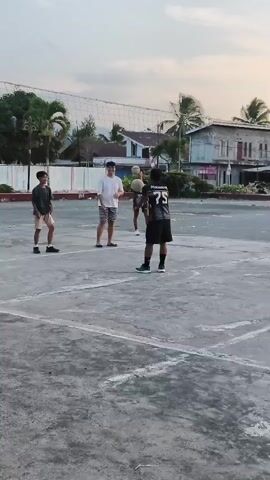 Four young males are playing volleyball on a paved court with white lines. Two are on the left side of the net, and two are on the right. The sky is overcast, and palm trees and buildings are visible in the background. One male on the right side of the net is jumping to hit the ball over the net. Another male on the left side of the net is also in motion, likely preparing to receive the ball. One male on the right is wearing a black and white jersey with the number 75.
