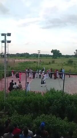 The video captures an outdoor gathering, possibly a celebration or cultural event, taking place on a fenced-in court area. A group of people, many wearing white robes or traditional attire, are engaged in some form of organized activity or dance on the court. Some individuals in the foreground appear to be observing the event. The environment suggests a sports facility or an open field with a cloudy sky overhead. Lighting towers are visible, indicating the possibility of the event continuing into the evening. There are trees and vegetation surrounding the court, and a low-lying area with more foliage is visible in the foreground. The perspective is from a raised vantage point, looking down at the scene.