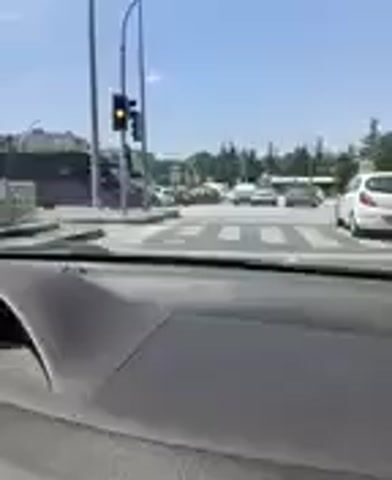 The video shows a view from inside a car, looking out through the windshield and side windows. The car appears to be stopped or moving slowly in traffic. Outside the car, there are other vehicles, including cars and a van. The environment appears to be an urban or suburban setting with buildings and trees visible in the background. The sky is clear and blue. There are traffic lights and what appears to be a crosswalk. A person's hand is visible on the steering wheel. The video is shaky, suggesting it was recorded by someone holding a phone or camera while in the moving vehicle.