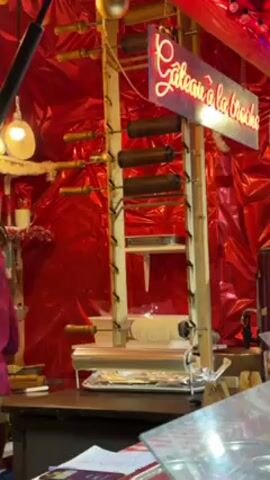 The video shows a "Gateau à la broche" stand, likely at a Christmas market, featuring a wooden structure with rotating cylinders. The stand has a neon sign that reads "Gateau à la broche". A woman in a pink top stands behind the counter, attending to a customer. The background is decorated with shiny red material.  A metal container and candy canes can be seen on the counter, along with what appears to be a cash register.