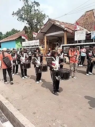 A marching band parades down a street. The band members are wearing matching uniforms. Some are carrying drums and drumsticks. The band is performing outdoors. The background shows a building with a red roof and trees. There are onlookers on the side of the street watching the parade. The overall atmosphere is festive.