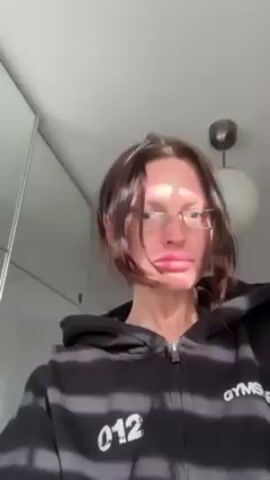 A woman with short brown hair and glasses is facing the camera. She is wearing a black zip-up sweatshirt that says "Gymshark" on the sleeve. She holds up her hand to show her nails, which are painted white. She mentions that she used to bite her nails, but now they look nice. The background includes white cabinets and a round light fixture on the ceiling.