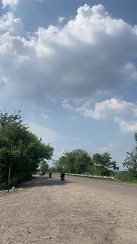The video captures a roadside scene on a sunny day with a blue sky dotted with puffy white clouds. The foreground features a dirt and gravel shoulder of the road, littered with trash. To the left, there is a thick growth of green trees. The main subject of the video is the paved road, where several motorcycles and a white car are traveling in the same direction. On the right side of the road, more trees are visible and there are also road signs.