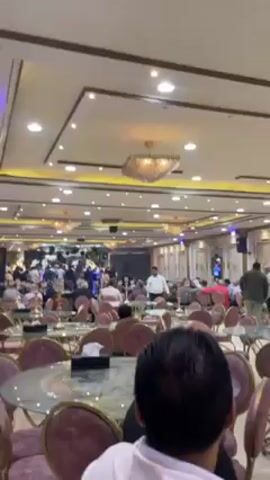 The video shows a lavishly decorated banquet hall with a large crowd of people, many seated at tables. The chairs are upholstered in a burgundy color with marble-topped tables. There's a stage at the far end of the room, where people are gathered and dancing, illuminated by stage lighting. The ceiling is decorated with recessed lighting and ornate chandeliers. The overall atmosphere appears celebratory, possibly a wedding reception or a similar social event.