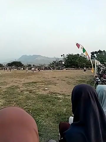 The video shows a soccer game being played on a dirt field. There are two teams of players, one in white and blue uniforms and the other in black uniforms. The game appears to be in progress, with players running and kicking the ball. There are spectators on the sidelines, many of whom are women wearing hijabs. There are also trees and buildings in the background, as well as a mountain in the distance. The sky is overcast. A string of colorful flags is visible on the right side of the frame, near a soccer goal. The camera is focused on the game from behind some spectators.