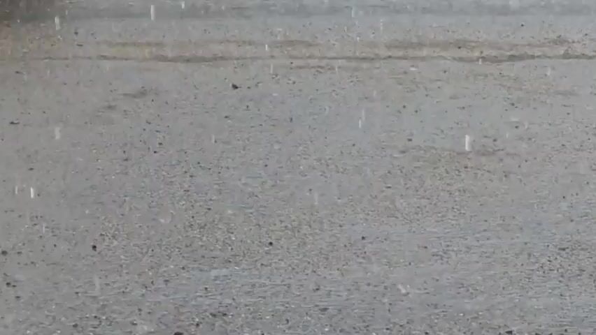 The video shows a close-up, slightly blurred view of raindrops falling onto a textured, grayish surface, possibly asphalt or concrete. The drops create small, transient splashes or ripples upon impact. The background is also out of focus and appears to be a continuation of the same surface. The lighting suggests an overcast or dim environment, consistent with rainy weather. No people or other objects are visible in the frame.