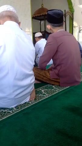 The video shows an interior view of what appears to be a mosque. Several men are seated on the green carpet, facing away from the camera, seemingly in prayer or contemplation. One man in the foreground is wearing a maroon robe and a black cap. Another man to his left is wearing a white robe and a white cap. In the background, a man in a white shirt and cap is standing and speaking into a microphone. The environment appears to be clean and well-lit, with light filtering in from windows. Decorative elements are visible, including a golden dome structure and ornate archways. The overall atmosphere suggests a religious gathering or service.