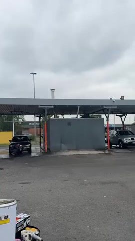 The scene shows an outdoor car wash on an overcast day. Two people are washing cars under a shared metal canopy structure. One individual is washing a white van on the left side of the frame with a water hose. A second person is positioned in front of a black sedan, seemingly attending to the front of the vehicle. A gray concrete structure is visible on the right side of the frame, possibly housing equipment for the car wash. In the background, across a parking area, is a McDonald's restaurant. In the foreground, there is a white drum-like container, and various car cleaning supplies. The ground is paved with asphalt and is damp from the ongoing car washing activities.