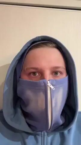 A person wearing a light blue hooded sweatshirt with the hood up and a face mask is centered in the frame. The face mask is also light blue and has a zipper down the middle. The person's eyes are visible, and they appear to be looking down slightly. The person's skin appears slightly flushed. The background is a plain white wall with a white door frame.