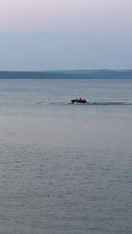 A small, inflatable boat is moving across a calm body of water. The boat is occupied by at least two people, both wearing orange life vests. The boat is leaving a visible wake behind it. The background consists of a hazy, tree-covered shoreline and a muted sky. The overall lighting suggests either dawn or dusk.
