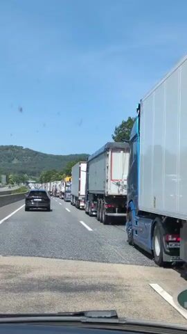 The video shows a traffic jam on a highway. Multiple trucks are lined up in lanes, moving very slowly or stopped. A black car is visible in the left lane, also moving slowly. The sky is clear and blue, with some trees and a hill in the background. The road surface is asphalt, with clear white lane markings. The perspective appears to be from inside another vehicle, looking forward. The trucks are of various types, including semi-trailers and tankers. Some trucks have company logos or text visible, such as "BERG SCHNEIDER" and "SCHMIDT". The overall impression is of a congested highway on a sunny day.