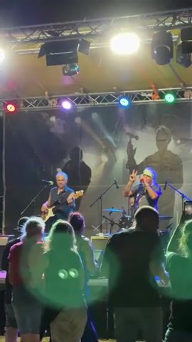 A live band performs on an outdoor stage at night. The stage is lit by spotlights and colored lights. A banner with shadowy figures is displayed behind the band. In the foreground, a crowd of people watches the performance. The band consists of at least two members. One musician, on the left, plays a white bass guitar and wears a black sleeveless shirt. The other musician, on the right, sings into a microphone while wearing a purple shirt and a white bandana. He holds up two fingers in a peace sign. The crowd is diverse in age and gender. Some members of the audience are holding up their phones to record the concert. The overall atmosphere appears to be energetic and festive.