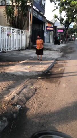 A woman wearing a floral shirt and a dark skirt stands on a sidewalk next to a white metal fence. The sun is shining, casting shadows on the ground. The sidewalk is made of brick and concrete. A street runs alongside the sidewalk, and a few buildings are visible in the background, including one with a sign that says "APOTEK." Another person, a woman wearing a headscarf and patterned pants, walks a bicycle past the first woman. The video appears to be taken from a moving vehicle.