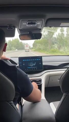 The video shows the interior of a car driving on a highway. The camera is positioned behind the driver, who is seen from the back wearing a dark blue t-shirt and with short dark hair. The driver's hands are on the steering wheel, and their right hand is near the center console where there is a touchscreen display. The touchscreen shows what appears to be climate control settings with diagrams of airflow and temperature adjustments. In front of the car, on the highway, a white truck with a red and white logo on its side is visible in the distance. The highway is bordered by green trees and vegetation. The sky appears overcast.
