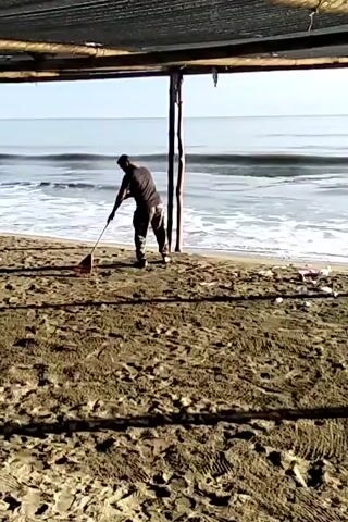 Here's a concise description of the video: 

A man is sweeping a sandy beach with a rake. He is silhouetted against the bright daylight and ocean waves. The view is framed by the structure of a covered shelter with a partially visible ladder to the left. The beach sand is dark and textured.