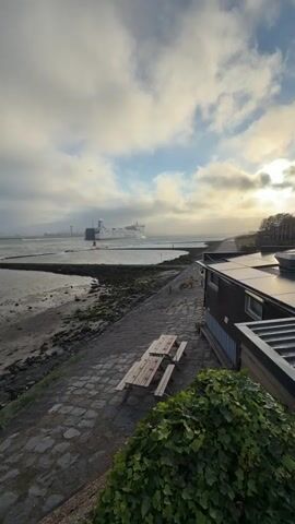 This video shows a scenic view of a body of water, likely the sea, with a ferry in the distance. The foreground features a paved walkway with picnic tables and a building with solar panels. The beach area to the left is rocky and covered with some seaweed. The sky is cloudy with patches of sunlight breaking through, suggesting it's either sunrise or sunset. The overall atmosphere is calm and serene.