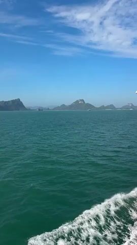 The video shows a view of the ocean from a boat moving through the water. The water is a deep turquoise color and shows a wake of white foam behind the boat on the right side. In the distance, a chain of islands with lush green foliage can be seen against a clear blue sky with scattered white clouds. The lighting suggests it is daytime, likely mid-morning or afternoon. The overall scene is serene and appears to be a scenic view of a coastal or island landscape.