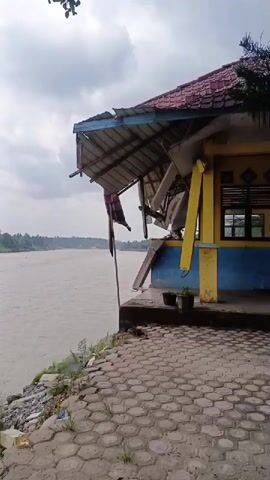 The video displays a damaged building situated on the bank of a wide, muddy river under an overcast sky. The building has a red tiled roof and brightly painted yellow and blue walls. Its front section, appearing to be a porch or canopy, is significantly damaged; the roof structure is bent, broken, and partially collapsed, with metal and wooden debris hanging down. A long, thin pole leans against the damaged structure, and a piece of fabric or flag-like material hangs from it. Two potted plants sit on the paved area in front of the building. The ground leading to the river is paved with hexagonal blocks, showing some small weeds growing through the cracks. Beyond the paved area, there are stones and some trash near the water's edge. The river itself appears calm but high, suggesting recent or ongoing water levels. In the distance, the river stretches towards a horizon lined with trees or vegetation. The scene depicts a state of structural damage, possibly due to erosion or an event impacting the riverbank, in Simpangjaya, Indonesia.