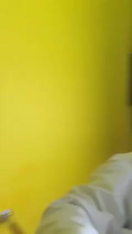 The video, shot from a low angle, shows a young woman with her hair in a braid, wearing a white long-sleeved shirt. She is looking upwards, presumably at the camera. The background consists of a high ceiling with a black and yellow color scheme. The ceiling is lined with what appear to be rectangular structures. There are bright fluorescent lights in the ceiling that create a strong glare. The room is brightly lit. There is some indistinct speech in the background.