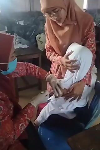 Two individuals in hijabs are administering what appears to be an injection to a child, also wearing a hijab and a white shirt. One individual, on the left, wears a face mask and gloves and holds a syringe. The other individual, on the right, holds the child and provides support. The setting appears to be an indoor space, possibly a classroom or a clinic, with tables and chairs visible. Other individuals in similar attire are seen in the background. The scene suggests a medical procedure, likely a vaccination, is taking place.