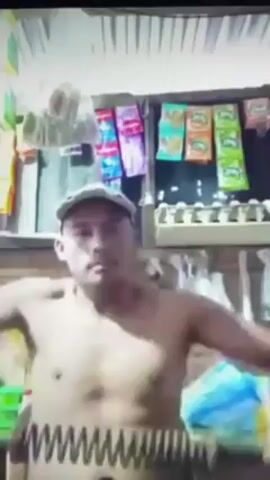A man with a light complexion, wearing a light-colored baseball cap, stands shirtless facing the camera. He moves his arms rhythmically in front of him. The background shows a small retail space with packaged snacks hanging on display behind him and an egg carton visible on a shelf. The lighting is dim.
