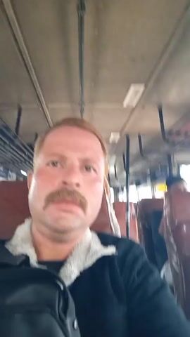 The video shows a person with fair skin, a mustache, and light brown hair riding on a bus. They are wearing a black jacket with a light-colored fuzzy collar and a black backpack slung over their shoulder. Other passengers are visible on the bus. The bus has red leather seats and advertisements on the ceiling. The view from the window is blurry, suggesting the bus is in motion. The camera is held by the person in the foreground, creating a selfie-style shot.