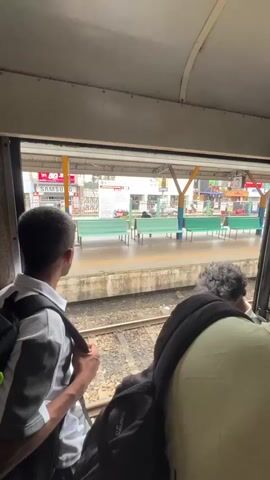 A short video shows the view from inside a train compartment, looking out a window at a train station platform. Two young men are visible from behind, seated in the train. The man on the left is wearing a white and gray striped shirt and a backpack. The man on the right has curly dark hair and is wearing a black backpack. The platform outside is lined with green benches and a covered waiting area with yellow and blue supports. Shops are visible in the background. Train tracks are in the foreground, and there is some debris on the tracks.