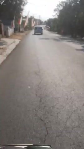 This is a video of a road scene. There are several cars and a motorcycle on the road. There are also some trees and flags on the side of the road. The sky is blue and there are some clouds. The video appears to be taken from a moving vehicle.