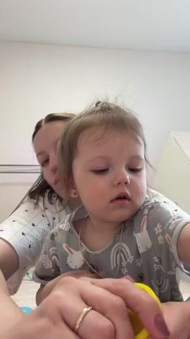A young woman and toddler are sitting indoors. The woman is behind the toddler and holding a small container of yellow Play-Doh, while the toddler holds a lump of blue Play-Doh. The woman is wearing a white shirt with a pattern and the toddler is wearing a gray shirt with a bunny pattern and a small necklace. The woman is holding the yellow container above her hand.