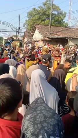 The video shows a street scene with a crowd of people gathered to watch a parade or performance. In the background, there are several individuals dressed in elaborate, colorful costumes, some wearing tall headdresses. They appear to be part of a procession or exhibition, with decorative elements and props visible. The foreground is filled with spectators, many of whom are wearing headscarves, looking towards the performers. Some individuals are holding up phones, likely to record the event. The setting appears to be a village or town, with houses and trees lining the street. The overall atmosphere is lively and festive.