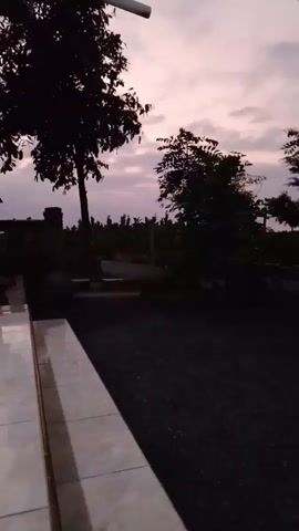 The video shows a dark outdoor scene during twilight. In the foreground, a tiled walkway runs along the bottom left of the frame. To the right of the walkway is a dark, gravelly surface. In the midground, silhouetted trees and vegetation are visible. The sky above is a soft pink and grey, indicating the time of day. A single person in dark clothing is partially visible walking away from the camera on the left side of the frame, near a building. The overall atmosphere is quiet and dusky.