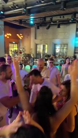 The video shows a group of people dancing in what appears to be a wedding reception or party. The environment is an indoor venue with a high ceiling, exposed lighting rigs, and large windows. The guests are dressed in light-colored or white clothing, with some women in dresses and men in shirts and pants. There are tables and chairs visible in the background, suggesting a dining area or seating arrangements. The foreground shows several individuals in close proximity, actively dancing and interacting. A videographer with a camera is also present, documenting the event. The atmosphere appears festive and energetic.
