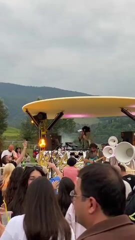 The video shows an outdoor music festival or gathering. A crowd of people is visible, many dancing and holding up their hands. A stage with lights and speakers is set up in the background, with musicians or DJs performing. The environment appears to be a grassy field with trees and hills in the distance. The sky is overcast. People are dressed casually in summer clothing. Some individuals are holding drinks or phones. The overall atmosphere seems festive and energetic.