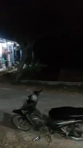 A black motorcycle is parked on a dirt road at night. A house is visible in the background, with lights on inside. There are people inside the house, but their features are indistinct.