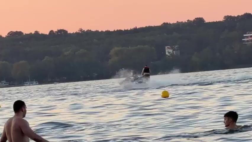 The video shows a body of water with several people in it and on jet skis. The sky is a pale orange, suggesting it is sunset or sunrise. In the foreground, there are at least three men in the water, two of whom are looking towards the right side of the frame. One man is facing away from the camera, and another is partially visible on the far right. In the middle ground, two jet skis are being ridden by individuals. The jet ski on the left is creating a significant spray of water. The jet ski on the right is further away and appears to be moving more slowly. In the background, a tree-covered hill slopes down to the water. Several boats are moored near the shore. There are also yellow buoys floating in the water. The overall impression is a leisurely scene at a lake or river.