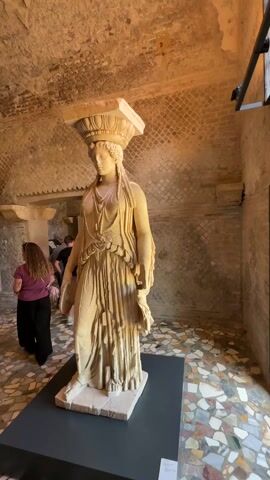 The video shows the interior of an ancient Roman building or museum. In the foreground, there are three Caryatid statues displayed on black platforms. Each statue depicts a woman in draped clothing, holding a flat, ornamented square on her head. The statues are made of light-colored stone or plaster. The background features a mosaic floor of various colors, and the walls are made of aged brick or stone. A woman in a pink top and black pants walks towards a white sign with writing on it in the background. Another person is also visible in the background. The ceiling is arched and made of similar material as the walls.  The lighting is a mix of natural light from an unseen source and artificial light from ceiling fixtures.