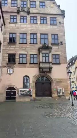 A medium shot shows the front of the Stadtmuseum Fembohaus in Nuremberg, Germany. The building has a light-colored stone facade with a pink lower level. There are rows of dark-framed windows on each floor. A decorative balcony protrudes from the second floor. The entrance has large double doors. A plaque with the number "5" is visible on the left side of the building. A lamppost stands to the right, and blurred figures of people under umbrellas are visible in the background. The ground is paved with cobblestones. The sky is overcast.