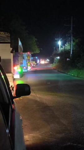 The video captures a night scene from inside a vehicle, looking out onto a road in Meuse, Indonesia. A line of trucks is visible ahead, their tail lights, marker lights (including green and blue), and headlights creating a vibrant glow against the dark environment. The asphalt road reflects these lights, suggesting a damp or glossy surface. To the right of the road, utility poles and trees are partially illuminated by a bright streetlight further down the road. The immediate foreground shows the side mirror of the viewer's vehicle. The vehicles appear to be stationary or moving very slowly, indicating traffic or a potential stop. No people are visible, and no discernible text is present in the frame.