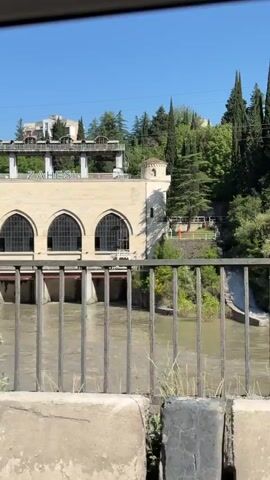 The video, shot from a moving vehicle, displays a large, beige-colored building with multiple arched windows and a prominent "ZAHES" sign on its upper facade. A wide, murky river flows in front of the building. A metal railing and a concrete barrier pass in the immediate foreground, while trees and greenery intermittently obscure the view. The background shows hills and a clear blue sky.