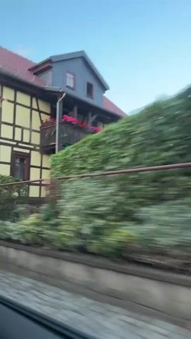 The video shows a fast-moving view from a vehicle, capturing glimpses of buildings and street scenes. The first shot focuses on a building with a yellow facade and dark timber framing, featuring several windows and a balcony adorned with pink flowers. A cobblestone pathway and a fence are visible in the foreground. The subsequent shots are blurred due to the motion, showing parts of buildings with stone walls, slate siding, and a red-painted exterior. The video appears to be taken while driving through a residential area, possibly in Europe, given the architectural style. The time is indicated as approximately 9:30 AM.