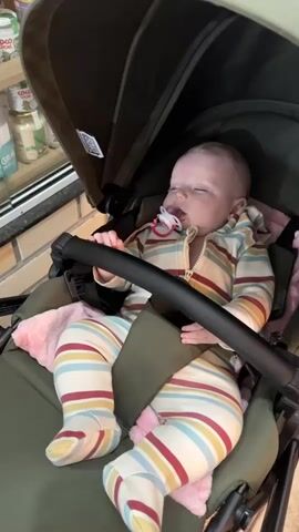 A baby is sleeping in a stroller. The baby is wearing a striped hooded jumpsuit and matching footed pants. The stroller is dark green and has a black handlebar. The baby has a pacifier in their mouth and is holding onto the handlebar with one hand. A pink blanket is underneath the baby. There are jars of coconut oil visible in the background.