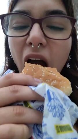 A close-up shot shows a young woman with glasses and a septum piercing taking a bite out of a hamburger. She is holding the hamburger with both hands, wrapped in white paper with blue and yellow text. Her eyes are closed as she chews. The background is blurred but appears to be outdoors with a building structure visible.