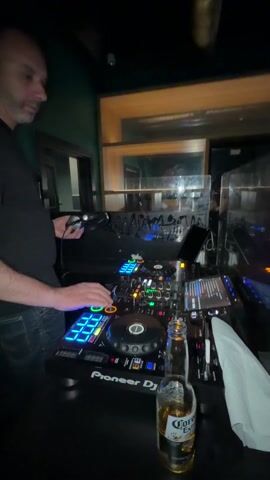 In a dimly lit setting, likely a bar or club in Grad Rijeka, Croatia, a male DJ is actively mixing music. He is seen from the waist up, wearing a black t-shirt with a "CK" logo and dark jeans. He stands behind an illuminated Pioneer DJ setup, which includes a mixer and turntables, all featuring various colorful lights that glow in the low light.

With both hands, he operates the controls, adjusting knobs and pressing buttons on the equipment. At one point, he briefly holds a small, dark object, possibly a microphone or a remote, to his mouth before placing it back down. To his right, a bottle, which appears to be a Corona beer, sits on the table alongside a white napkin.

The background features a dark green wall with a partial "BAR" sign visible on the left. Above, metallic pipes run across the ceiling. Behind the DJ, a wooden frame suggests a window or opening into another section of the venue. The overall atmosphere is dark and intimate, typical of a nightlife establishment. The DJ appears focused and engaged in his performance.