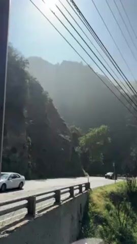 The video shows a road running alongside a mountain. The weather is sunny but slightly hazy. Several cars of different makes and colors are driving on the road, moving right to left. The road is lined with a low brick wall. Overhead are several electrical wires. The mountain is covered in trees and bushes.