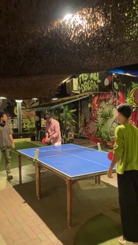 The video shows three individuals playing ping pong at an outdoor establishment at night. The ping pong table is blue with a white net. The people are dressed casually and are actively engaged in the game, with paddles in hand and the ball in motion. The background features graffiti art on the walls and a thatched roof overhead. The lighting suggests an evening or nighttime setting.