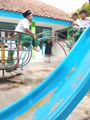 The video shows a school playground with children playing on different equipment. A blue slide is in the foreground, and further away, several boys wearing white shirts and green pants are on a merry-go-round. A girl in a purple skirt and white shoes is standing near the slide, and in the background, an adult wearing a black hijab is visible. Another boy, wearing a purple outfit and carrying a red backpack, walks toward a metal slide. The playground is tiled, and the buildings around have tiled roofs, suggesting a school environment.