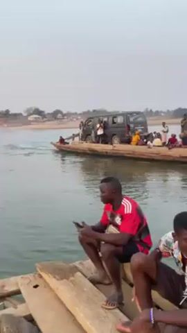The video shows a wide river scene. In the initial shot, two men are sitting on a makeshift wooden pier, one looking at his phone. In the background, a large, flat, wooden boat is ferrying a dark-colored van and several people across the river. The camera pans across the water, revealing a construction site in the middle of the river. The site contains sand, gravel, and heavy machinery including a crane and a pile driver. In the distance, there is a shoreline with trees. The sky is overcast.